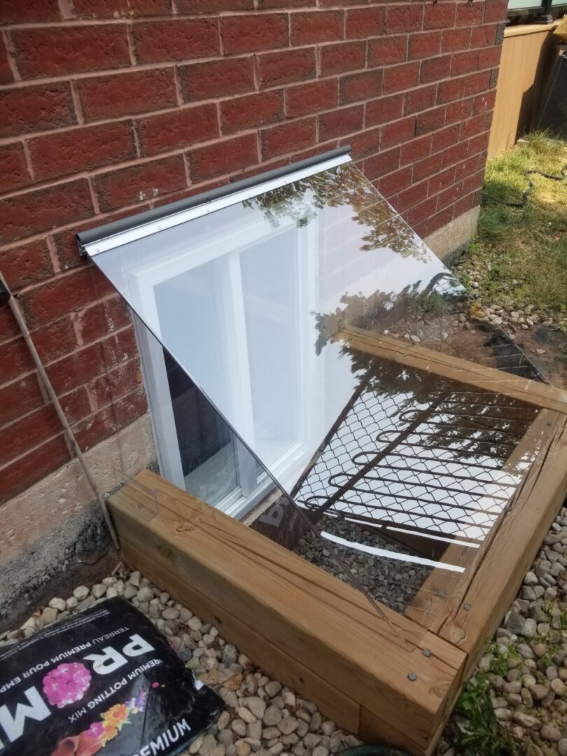 Slanted clear window well cover installed beside a Burlington-style brick home